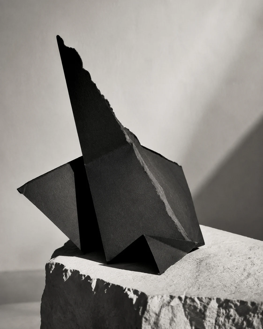 A torn folded black paper sculpture on pale limestone in directional light.