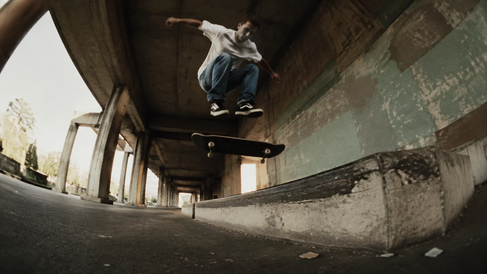 A skateboarder airborne beside a concrete ledge beneath a sunlit urban underpass.